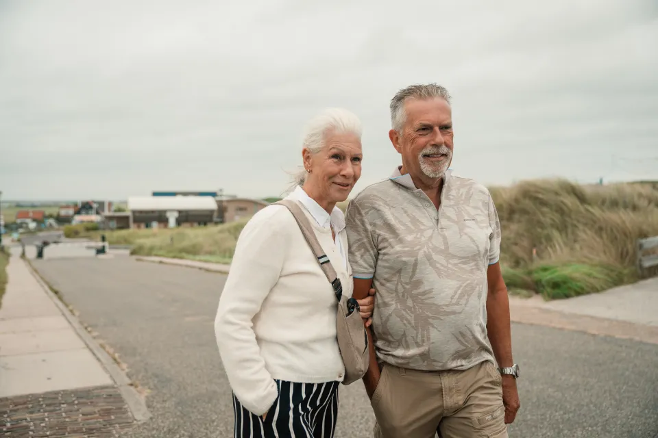 Ouder stel loopt lachend arm in arm over de strandweg bij de duinen